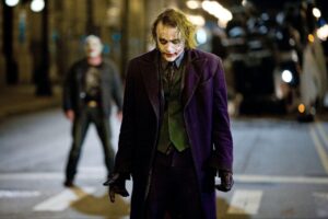 Кадр из художественного фильма The Dark Knight (2008): персонаж в клоунском гриме и фиолетовом пальто на городской улице, ночная сцена.